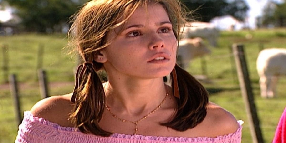 Íris em Laços de Família está vestindo uma blusa cor de rosa, usa o cabelo liso em duas partes amarrado e está ao ar livre em fazenda no fundo desfocada