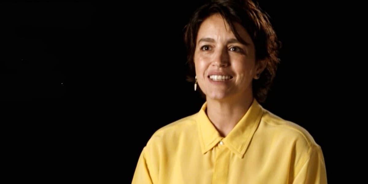 Manuela Dias, autora de Amor de Mãe, é uma mulher branca, de cabelos curtos castanhos usando uma blusa social amarela em um fundo preto