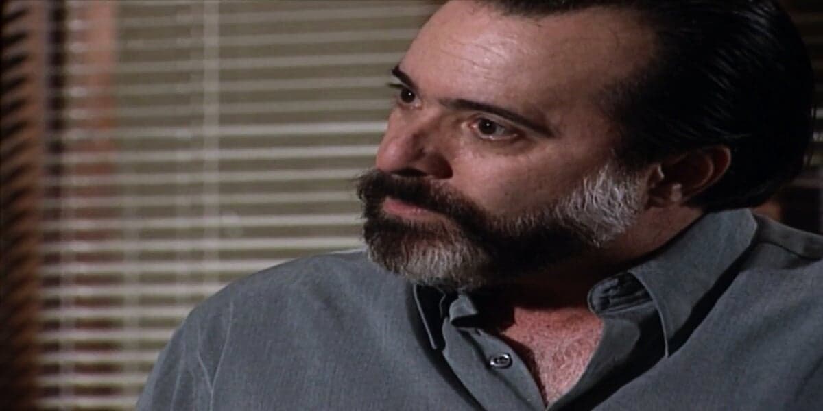 Miguel, homem que usa camisa social azul, é branco e tem barba da cor preta e branca está em cena da novela Laços de Família com expressão de raiva