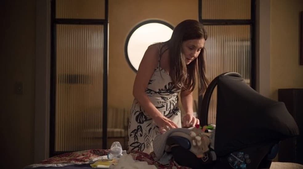 Thelma em cena de Amor de Mãe, mulher branca, com cabelos longos lisos e vestido florido está abaixada de frente ao bebê conforto em um quarto Thelma em cena de Amor de Mãe, mulher branca, com cabelos longos lisos e vestido florido está abaixada de frente ao bebê conforto em um quarto