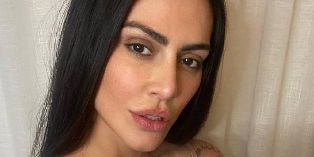 Cleo surge agachada, com a perna dobrada e arreganhada, coloca parte gigante na boca e fica com seios à mostra