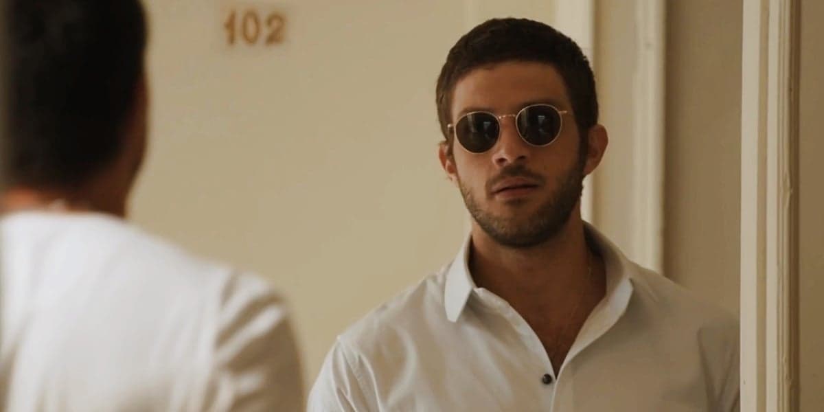 Ari usando camisa branca e óculos escuro parado em frente a porta aberta na novela Travessia Ari usando camisa branca e óculos escuro parado em frente a porta aberta na novela Travessia