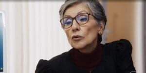Cidália é uma personagem de Travessia que usa blusa azul escuro, tem cabelos curtos grisalhos, usa óculos de grau marrom e é branca Cidália é uma personagem de Travessia que usa blusa azul escuro, tem cabelos curtos grisalhos, usa óculos de grau marrom e é branca