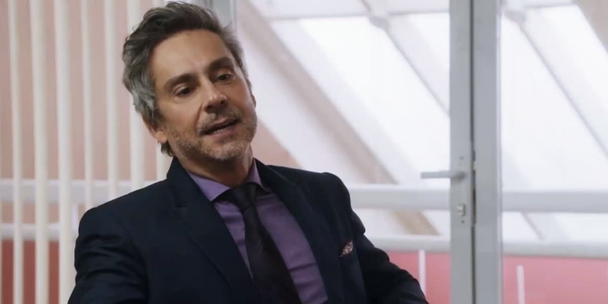 Stenio sentado em cena da novela Travessia, usando terno azul escuro, gravata vinho e camisa social roxa Stenio sentado em cena da novela Travessia, usando terno azul escuro, gravata vinho e camisa social roxa