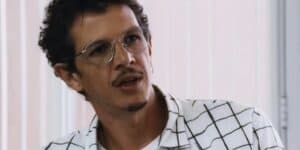 Gil em cena da novela Travessia usando camisa xadrez branca e preta, óculos de grau prata e cabelo curto com cavanhaque preto Gil em cena da novela Travessia usando camisa xadrez branca e preta, óculos de grau prata e cabelo curto com cavanhaque preto
