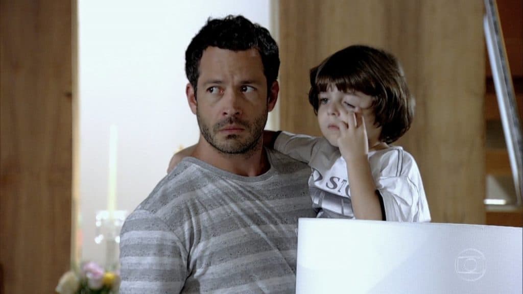 Quinzé com cara de desconfiado segura seu filho que coça o olho em cena da novela Fina Estampa Quinzé com cara de desconfiado segura seu filho que coça o olho em cena da novela Fina Estampa