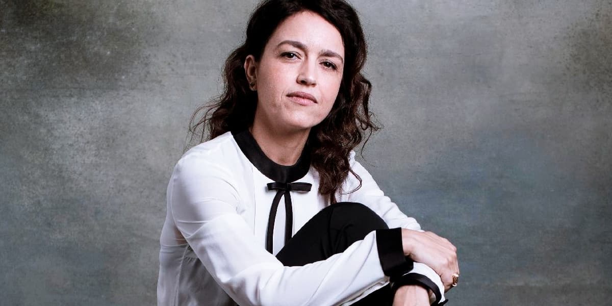 Manuela Dias sob um fundo cinza, é uma mulher branca, de cabelos cacheados castanhos que está sentada segurando sua perna com as mãos, usa roupas em tons brancos e pretos Manuela Dias sob um fundo cinza, é uma mulher branca, de cabelos cacheados castanhos que está sentada segurando sua perna com as mãos, usa roupas em tons brancos e pretos