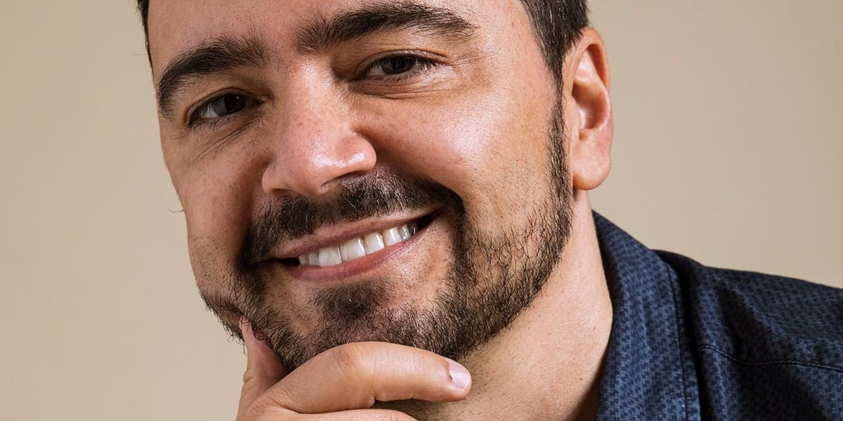 Rosto de Daniel Ortiz, autor de Salve-se Quem Puder, segurando o queixo e sorrindo ao usar uma camisa social azul Rosto de Daniel Ortiz, autor de Salve-se Quem Puder, segurando o queixo e sorrindo ao usar uma camisa social azul