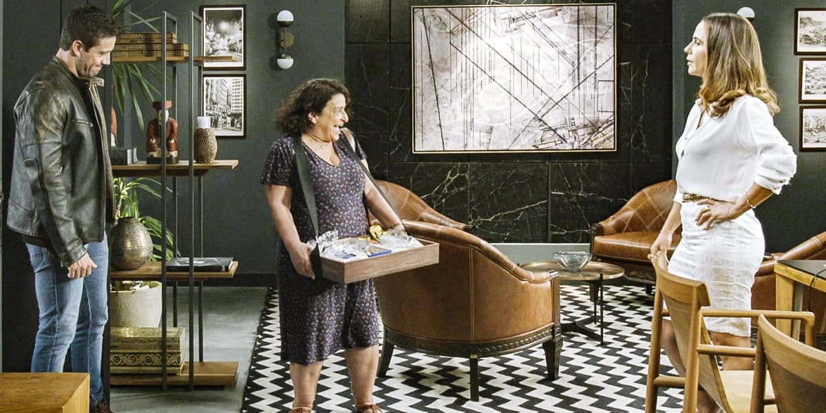 Ermelinda, usando vestido escuro e segurando caixa de madeira no meio de escritório de Dominique com vestido branco em cena de Salve-se Quem Puder Ermelinda, usando vestido escuro e segurando caixa de madeira no meio de escritório de Dominique com vestido branco em cena de Salve-se Quem Puder