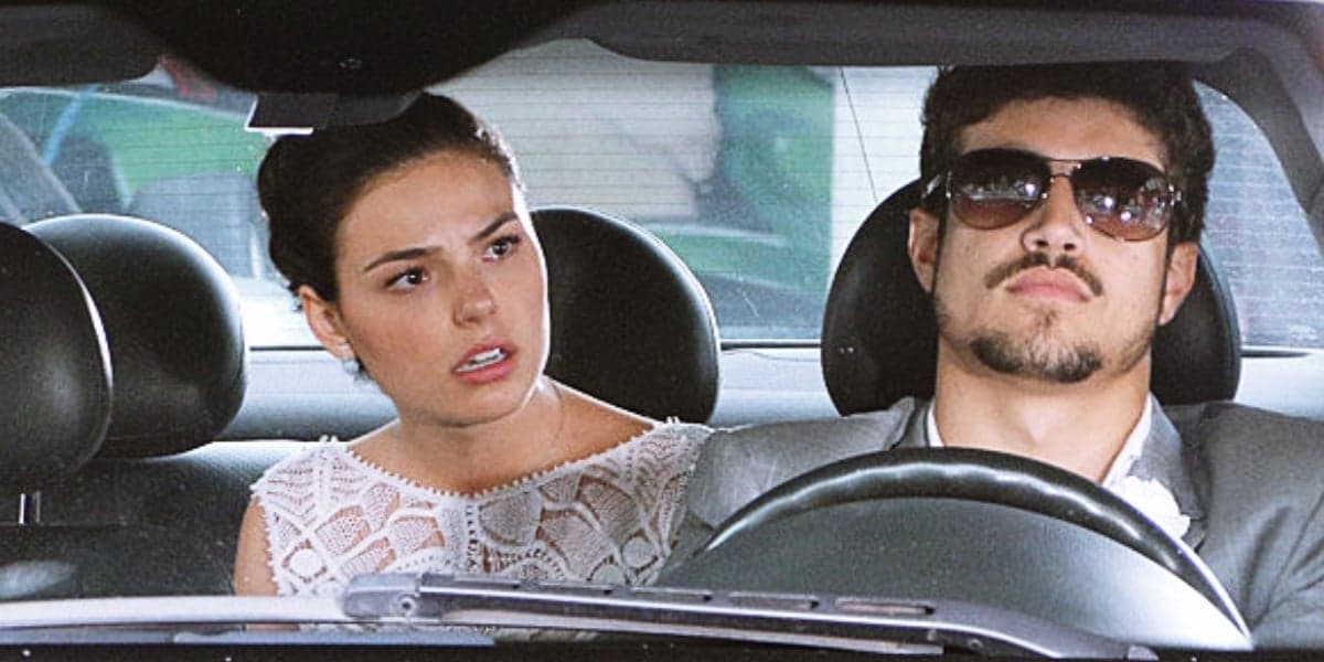 Marcela com expressão de surpresa com vestido branco está dentro de carro com Edgar que usa óculos marrom e terno cinza em cena da novela Ti-Ti-Ti Marcela com expressão de surpresa com vestido branco está dentro de carro com Edgar que usa óculos marrom e terno cinza em cena da novela Ti-Ti-Ti