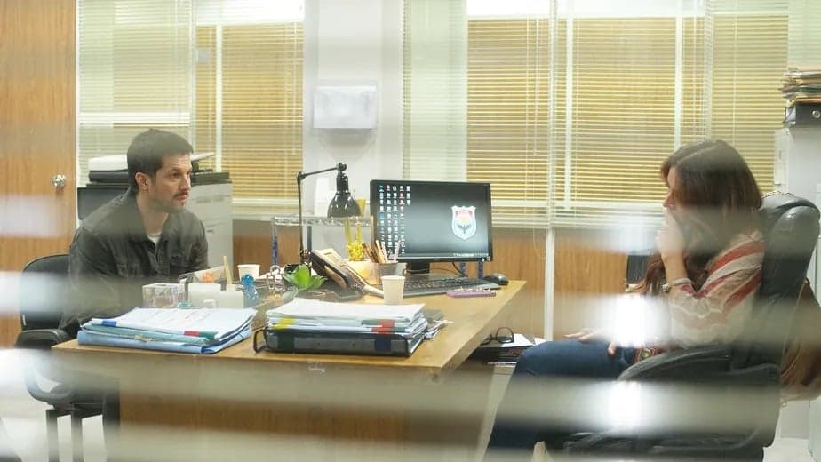 Oto e Helô estão em cena da novela Travessia dentro de uma delegacia sentados de frente um para o outro e separados por uma mesa Oto e Helô estão em cena da novela Travessia dentro de uma delegacia sentados de frente um para o outro e separados por uma mesa